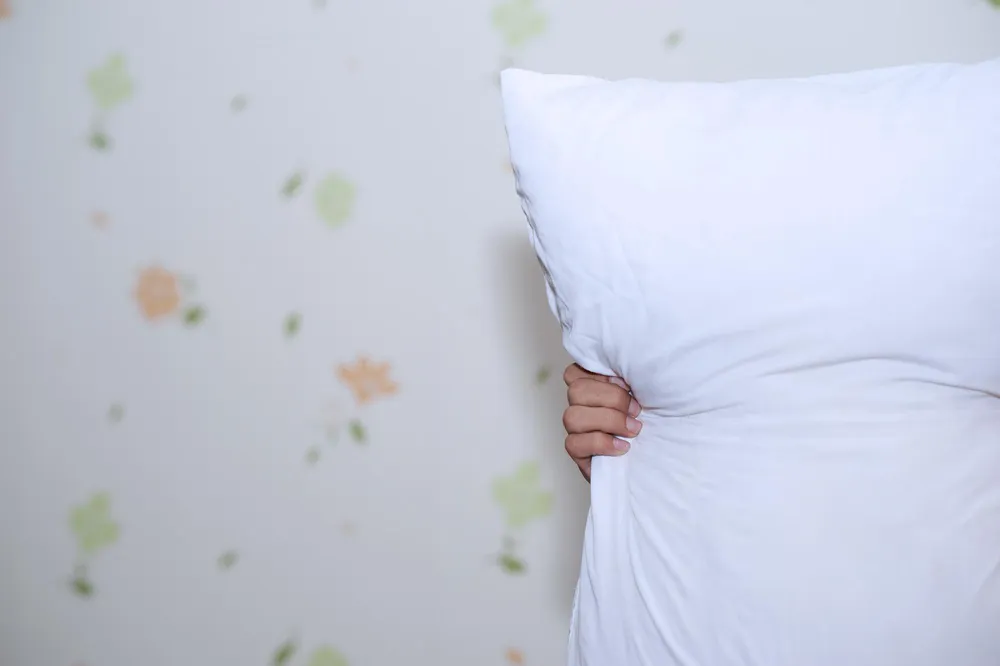 Close-up of an ergonomic memory foam pillow on a neatly made bed. A hand gently presses the surface, showing the slow-recovery foam and sculpted contour that cradles head and neck. Soft blur in the background highlights the pillow’s balanced blend of comfort and support, emphasizing healthy alignment rather than simple, flat softness.